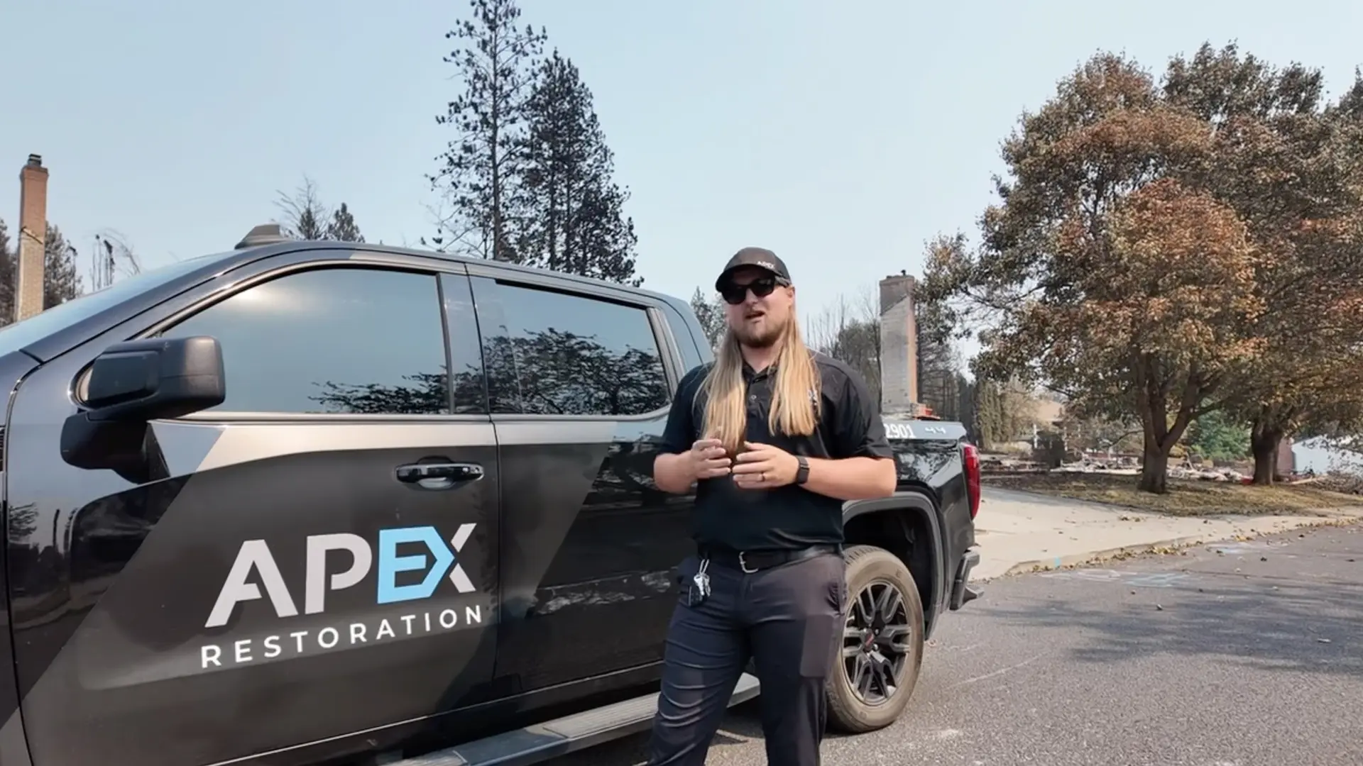 Crew member talking beside a branded pickup, with chimneys of burned homes behind, wildfire in Idaho and Eastern Washington